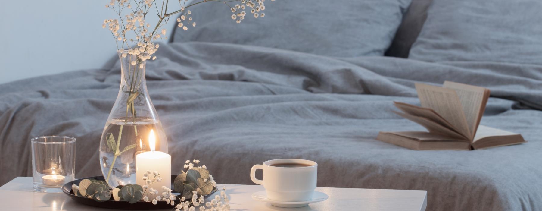 a bed with a denim gray comforter, coffee, candles, flowers, and a book